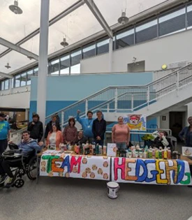 Group of students and staff standing proudly behind a colourful “Team Hedgehog” display table filled with items, inside a bright college atrium. Group of students and staff standing proudly behind a colourful “Team Hedgehog” display table filled with items, inside a bright college atrium.